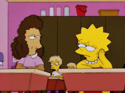 Two Simpson girls sit at classroom desks next to each other. One has blonde hair and one has brown hair. The one with brown hair blows bubblegum in the other's face.